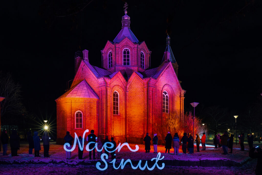 Punaisilla valoilla valaistu rakennus ja teksti "näen sinut"